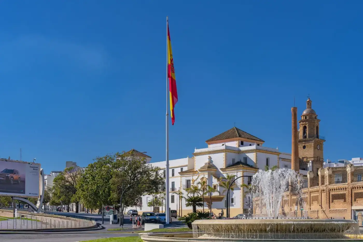 Scenic view of a plaza in Cádiz with a prominent Spanish flag, a fountain, and historic architecture.