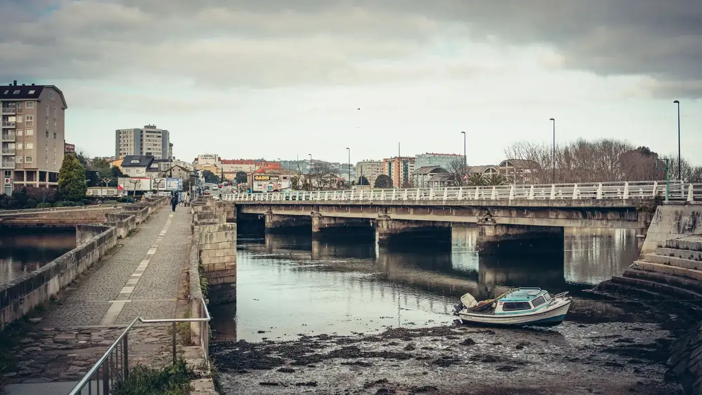 A peaceful urban landscape featuring a bridge over a river in Burgo, Spain, with buildings and a motorboat.