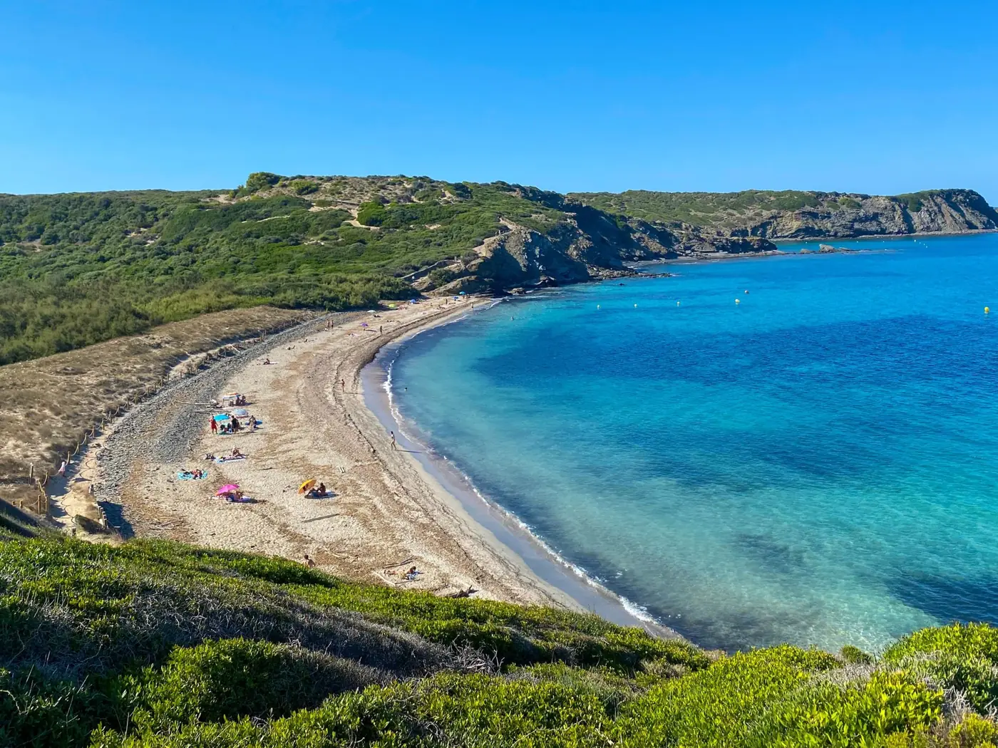 A stunning coastal view of a sandy beach in Ciudadela de Menorca, highlighting the clear blue sea and lush green hills.