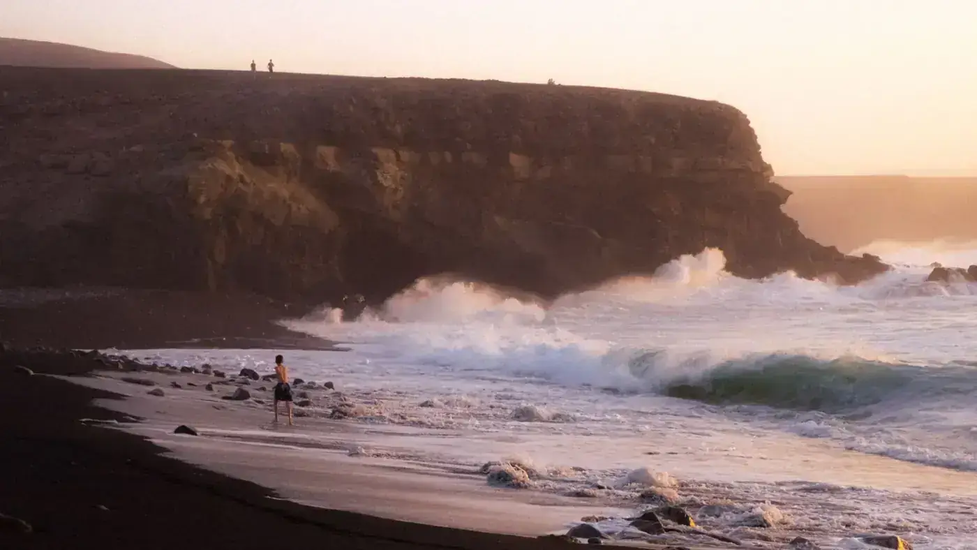 A lone person running along a scenic black sand beach in Spain with crashing waves and cliffs.