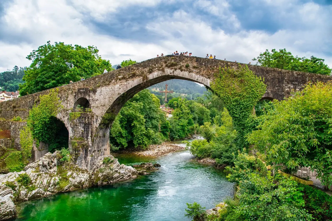 A picturesque ancient stone bridge arches over a lush green river, surrounded by vibrant trees and rich nature.