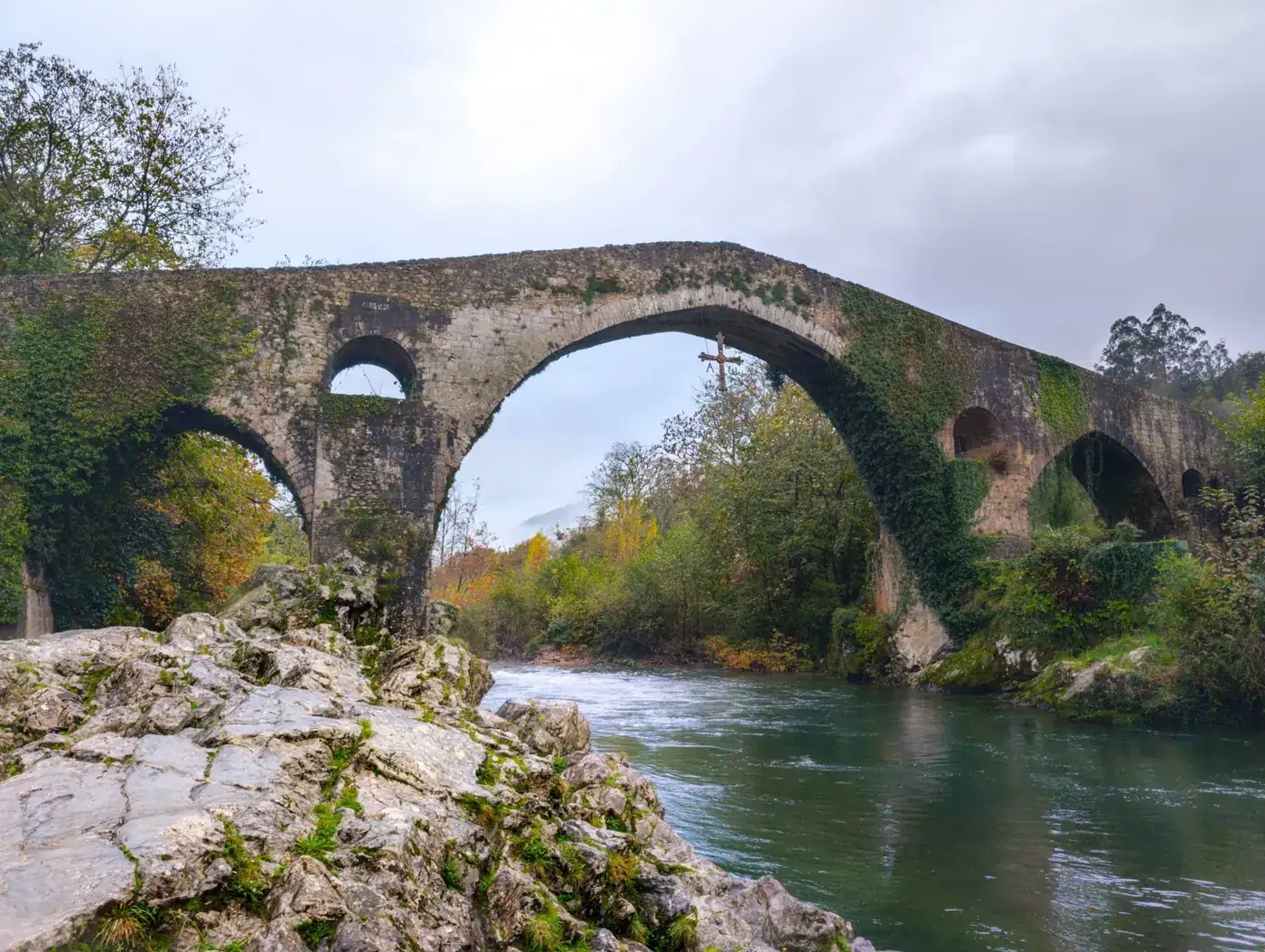 Scenic view of an ancient Roman stone bridge over the Sella River in Asturias, Spain, surrounded by lush autumn foliage.