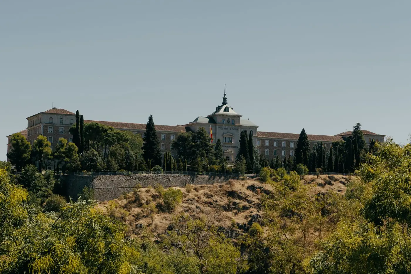 Historic Spanish Military Academy building in Toledo surrounded by trees under clear skies.