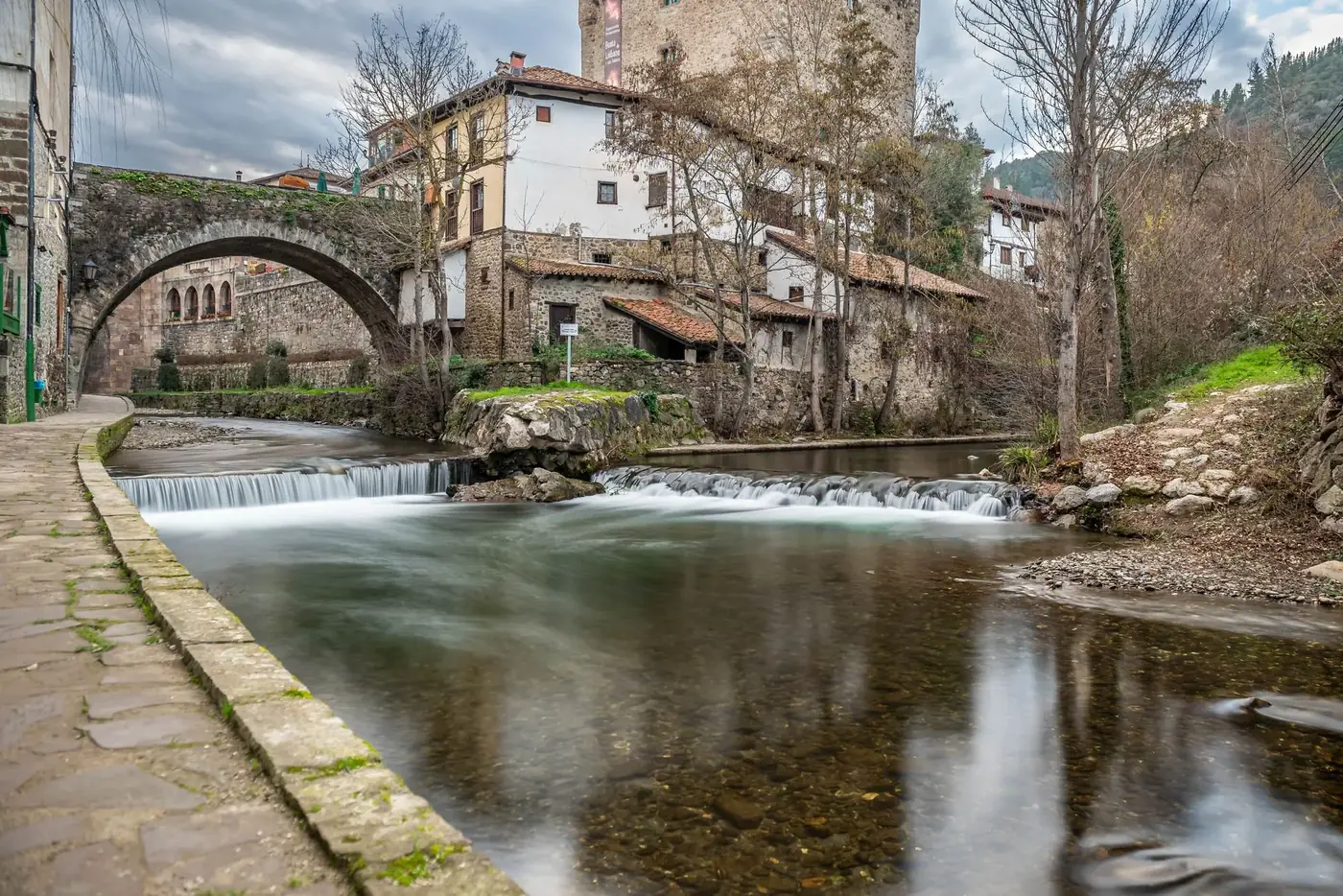 Tranquil river flowing under a stone bridge in a charming Spanish village, showcasing rustic architecture.