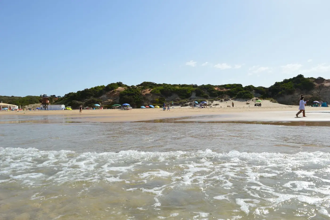 Capture of a sunny day at a wide sandy beach with distant sunbathers and colorful umbrellas.