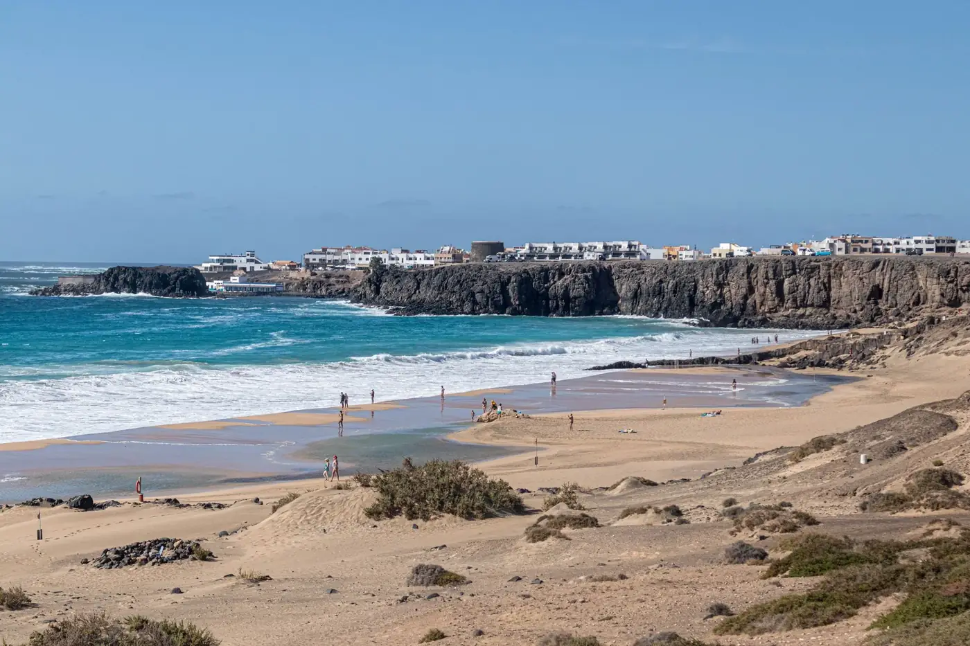 Scenic view of a sandy beach, cliffs, and coastal town under a clear blue sky.