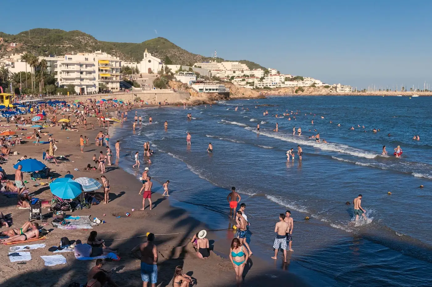 Lively scene of people enjoying a sunny day on the beach in Sitges, Spain.