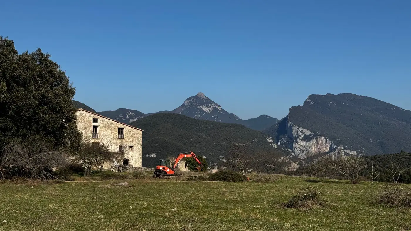 Charming stone house with excavator in scenic Catalan mountains under clear blue sky.
