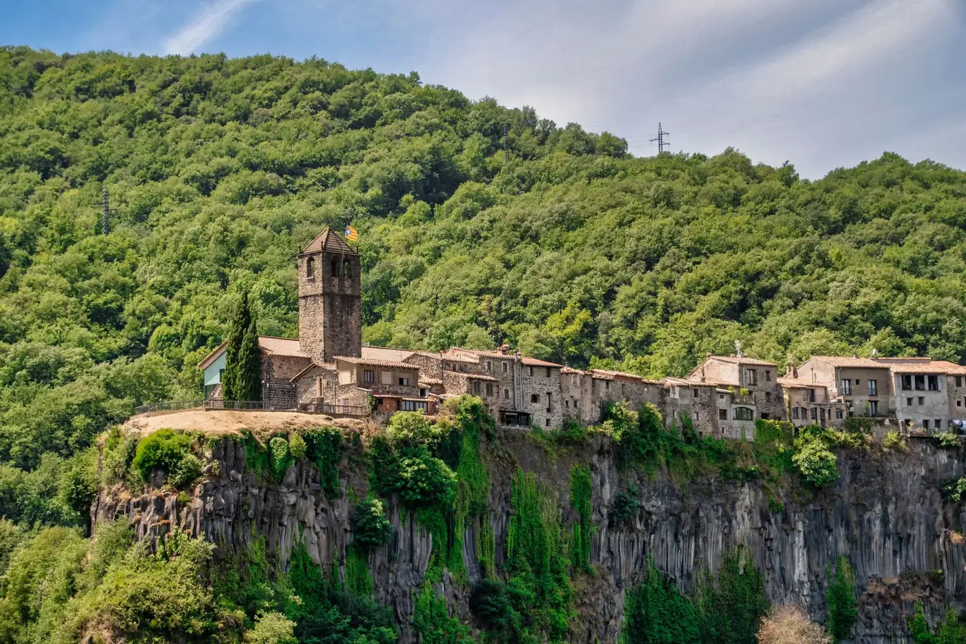 A scenic view of a medieval village perched on basalt cliffs with lush green forest in the background.