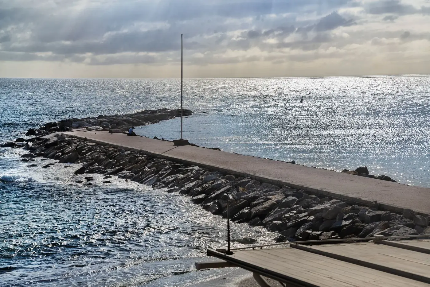 Calm beach scene with a paved jetty extending into the ocean under a cloudy sky.