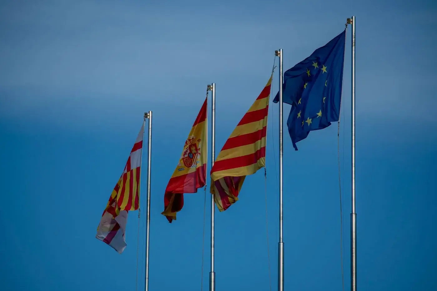 Flags of Spain, Catalonia, and Europe on flagpoles against a blue sky in Barcelona.
