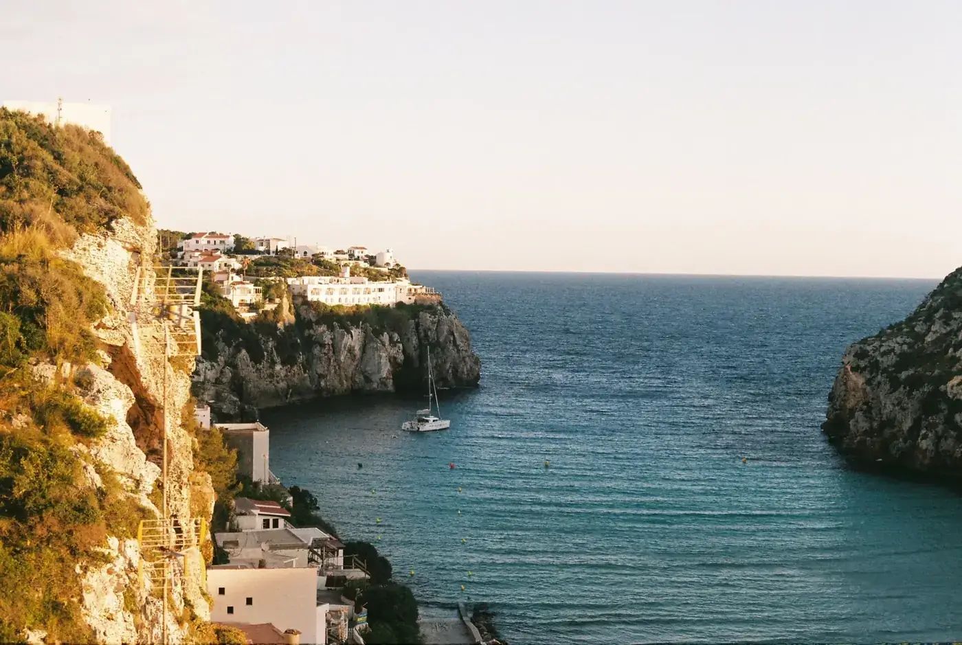 Dramatic cliffside view overlooking a serene bay with a yacht at sunset.