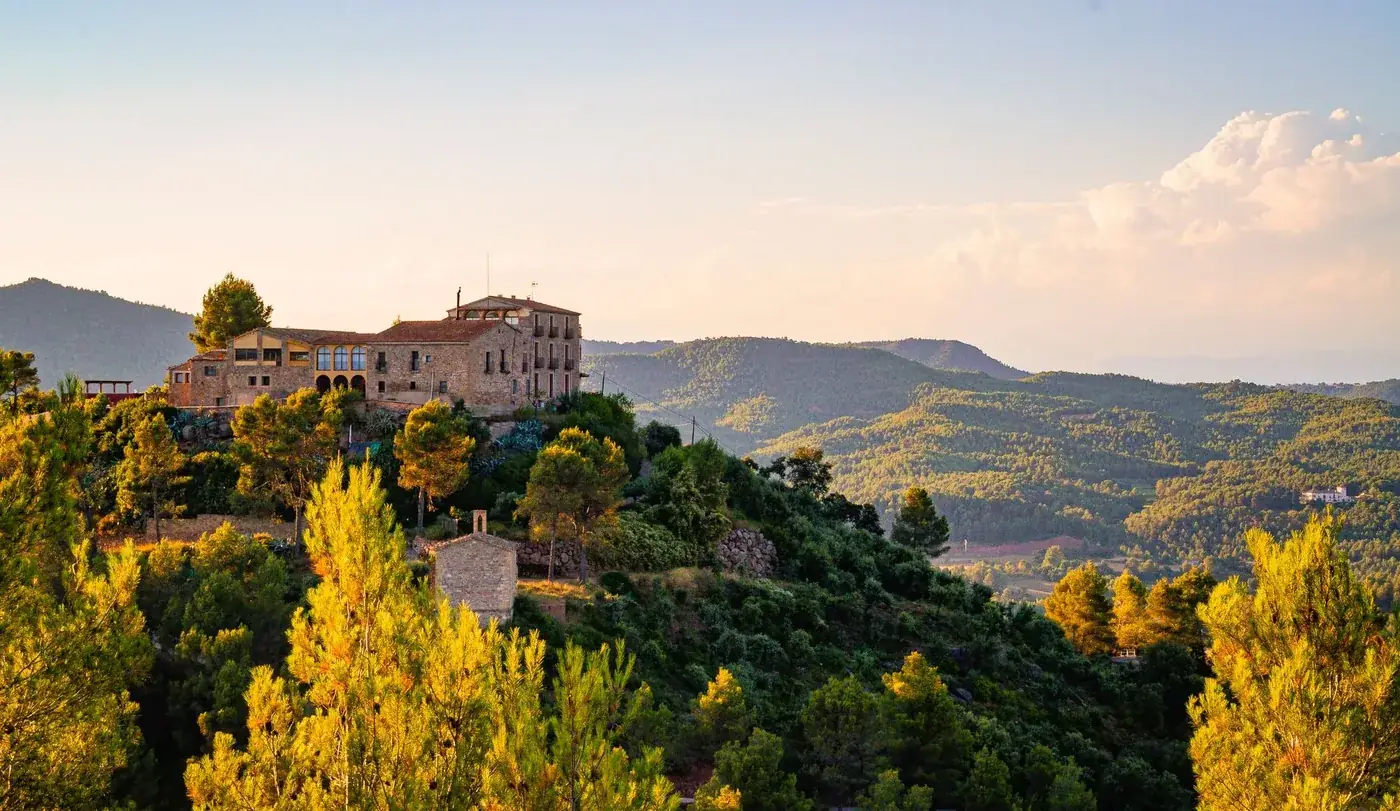 Historic building on a hillside in Catalonia surrounded by lush forests under a clear sky.