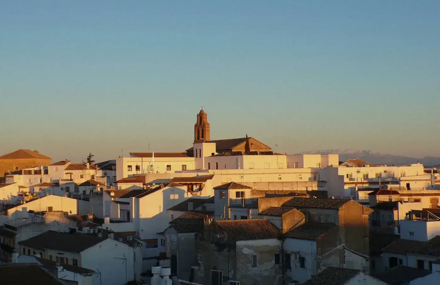A picturesque view of a Spanish village with white buildings at sunset, highlighting a church tower and distant mountains.