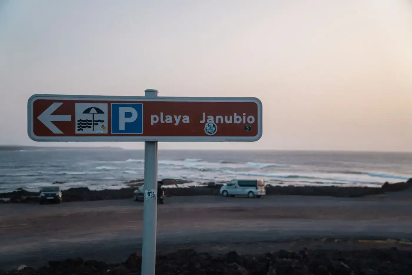 Beach sign at Playa Janubio pointing left under a pastel sky in Lanzarote, Spain with ocean in the background.
