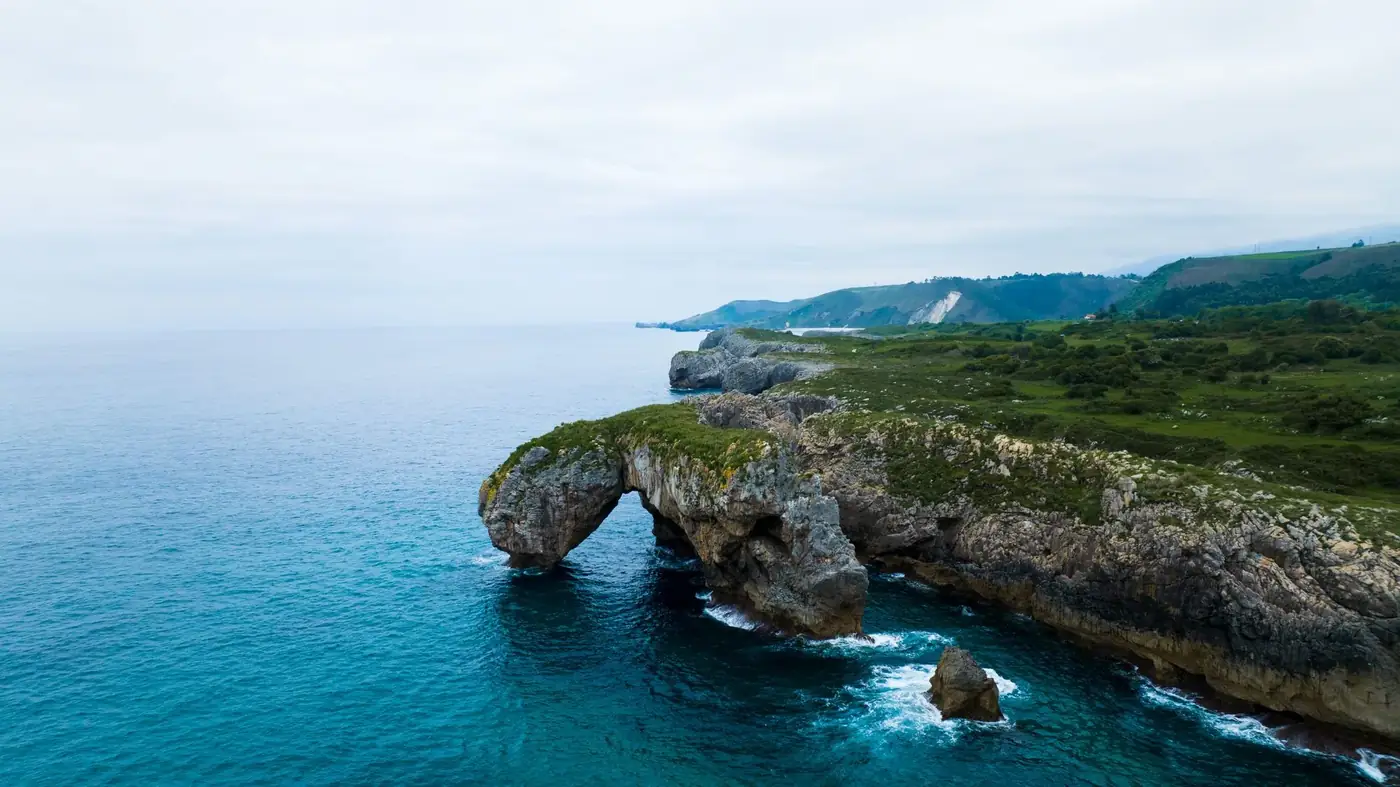 Stunning aerial view of the dramatic coastline at Asturias, Spain highlighting a rocky cliff arch and lush greenery.