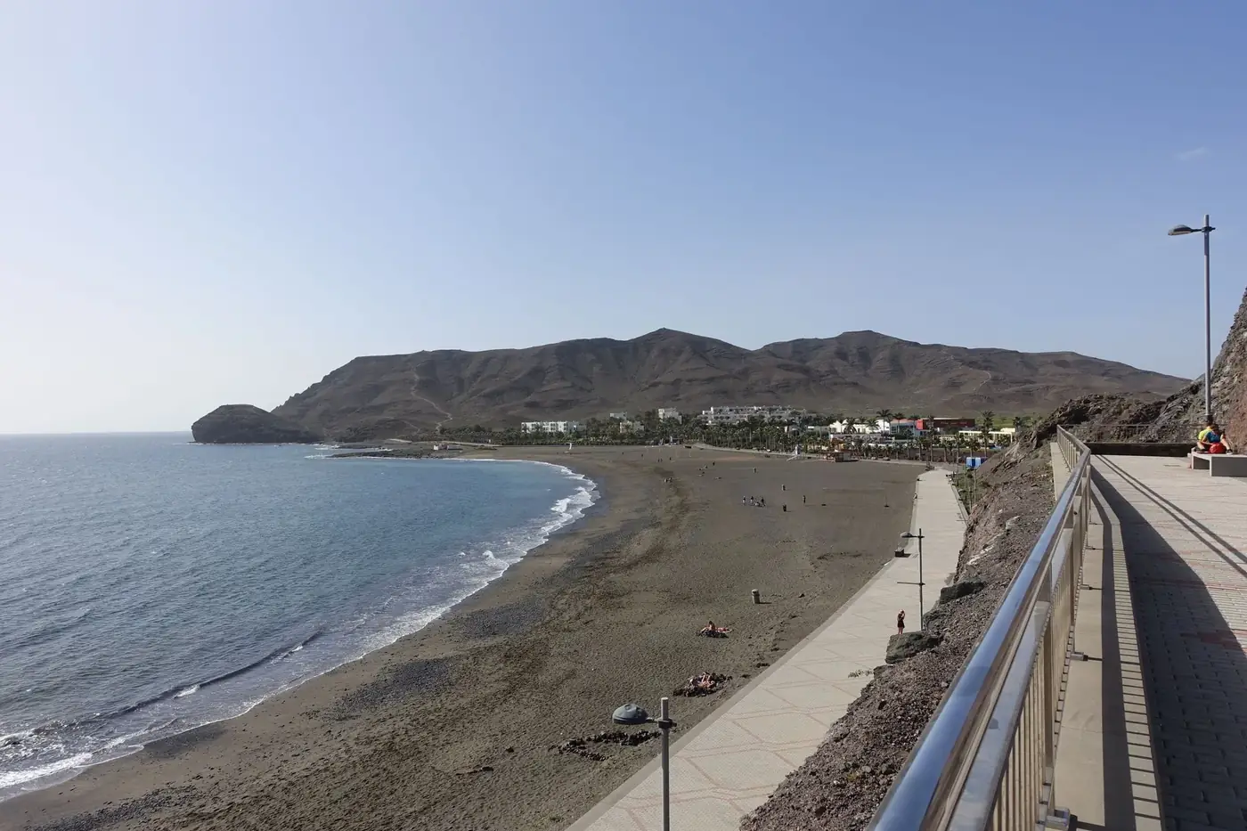 A peaceful beach with hills, promenade, and clear sky.