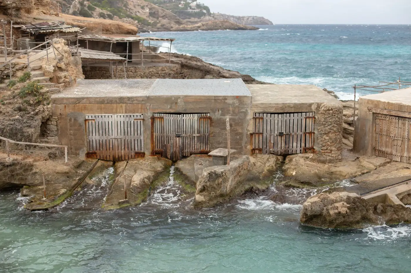 Rustic boathouses on Cala Tarida's Mediterranean coast in Ibiza, Spain, showcasing rocky terrain.