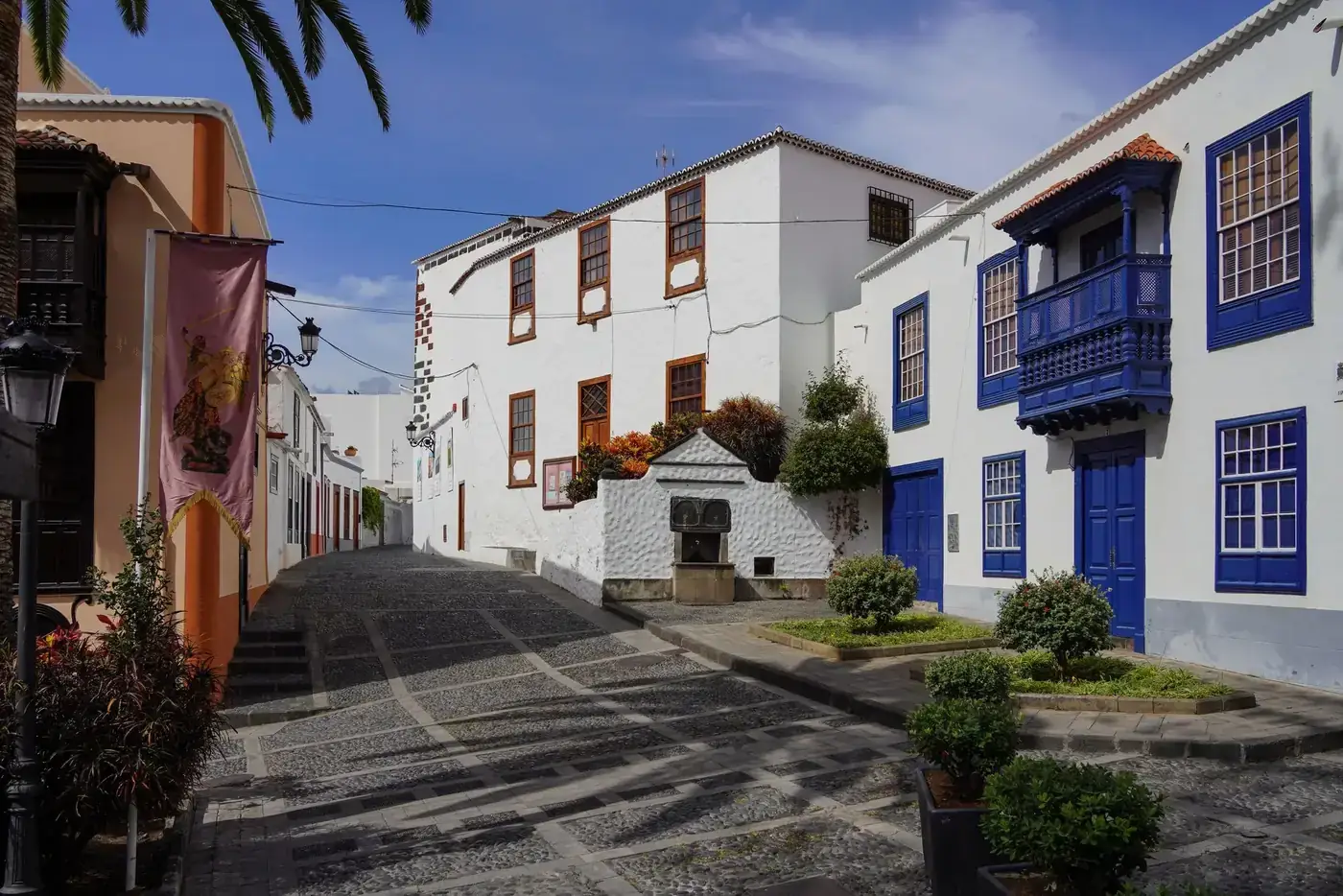 A picturesque cobblestone street in La Palma, Spain, with colorful houses and lush greenery under a bright blue sky.