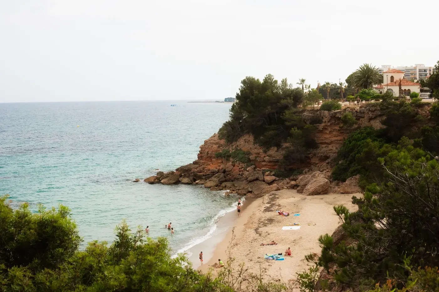 Peaceful sandy beach with rocky cliffs and tourists enjoying a sunny day by the coast.