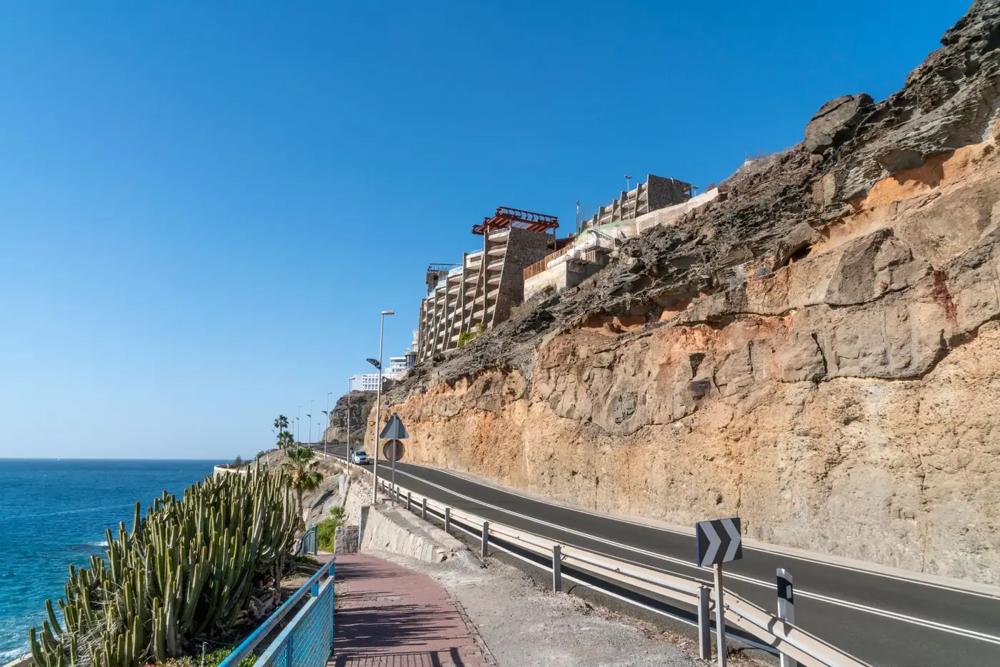 Empty asphalt road going between rocky cliff with residential buildings and calm sea against cloudless blue sky