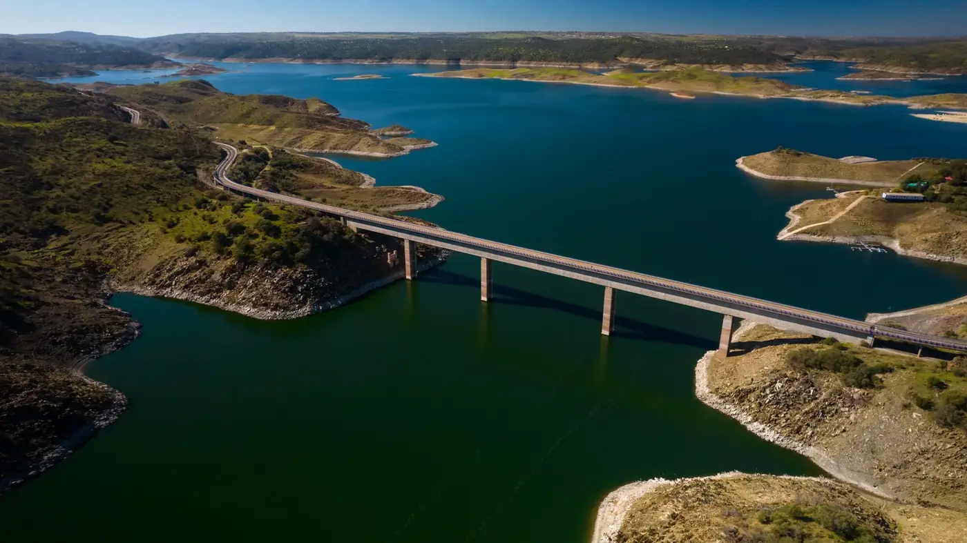 An aerial view captures a serene landscape with a bridge spanning over a calm reservoir in Cáceres, Spain.