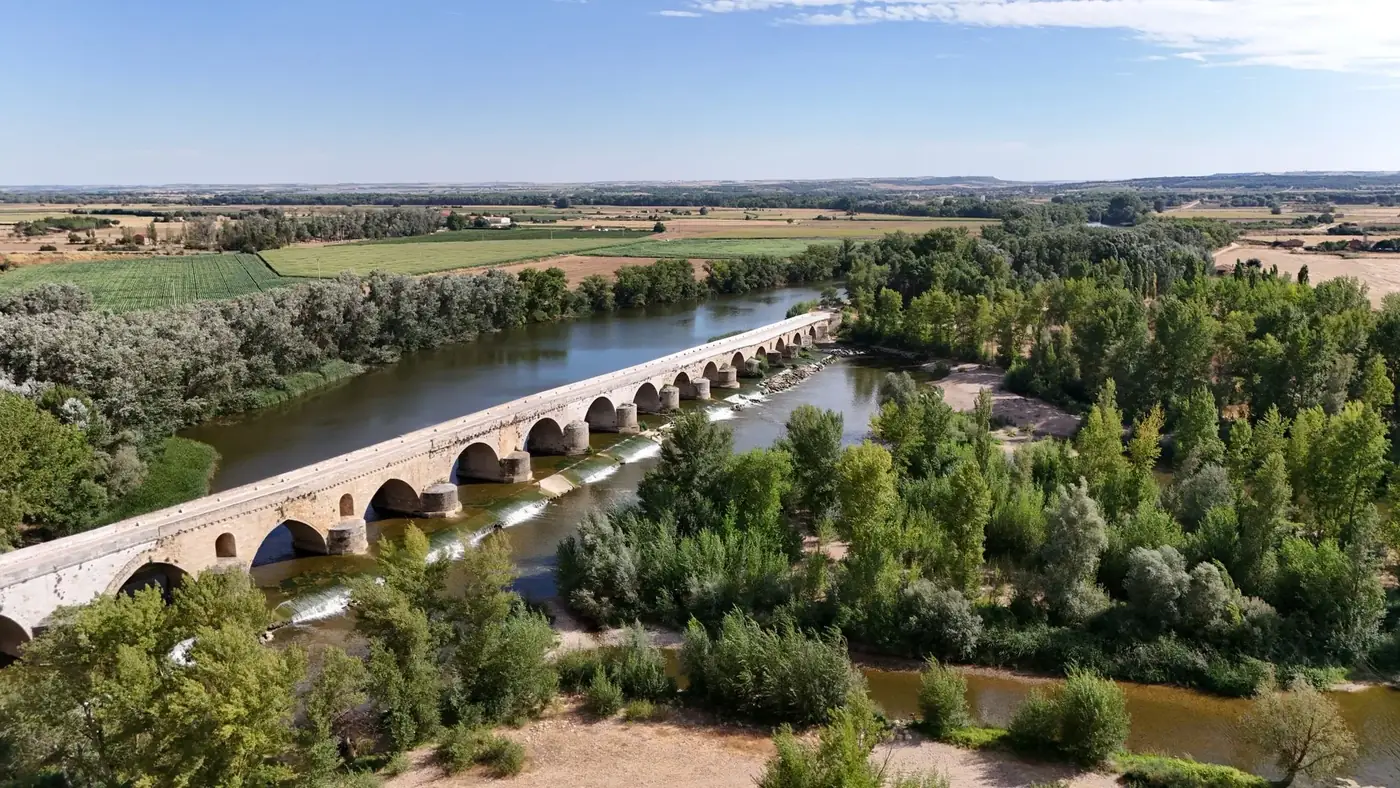 Aerial shot of a historic stone bridge surrounded by lush greenery and a river in Spain.