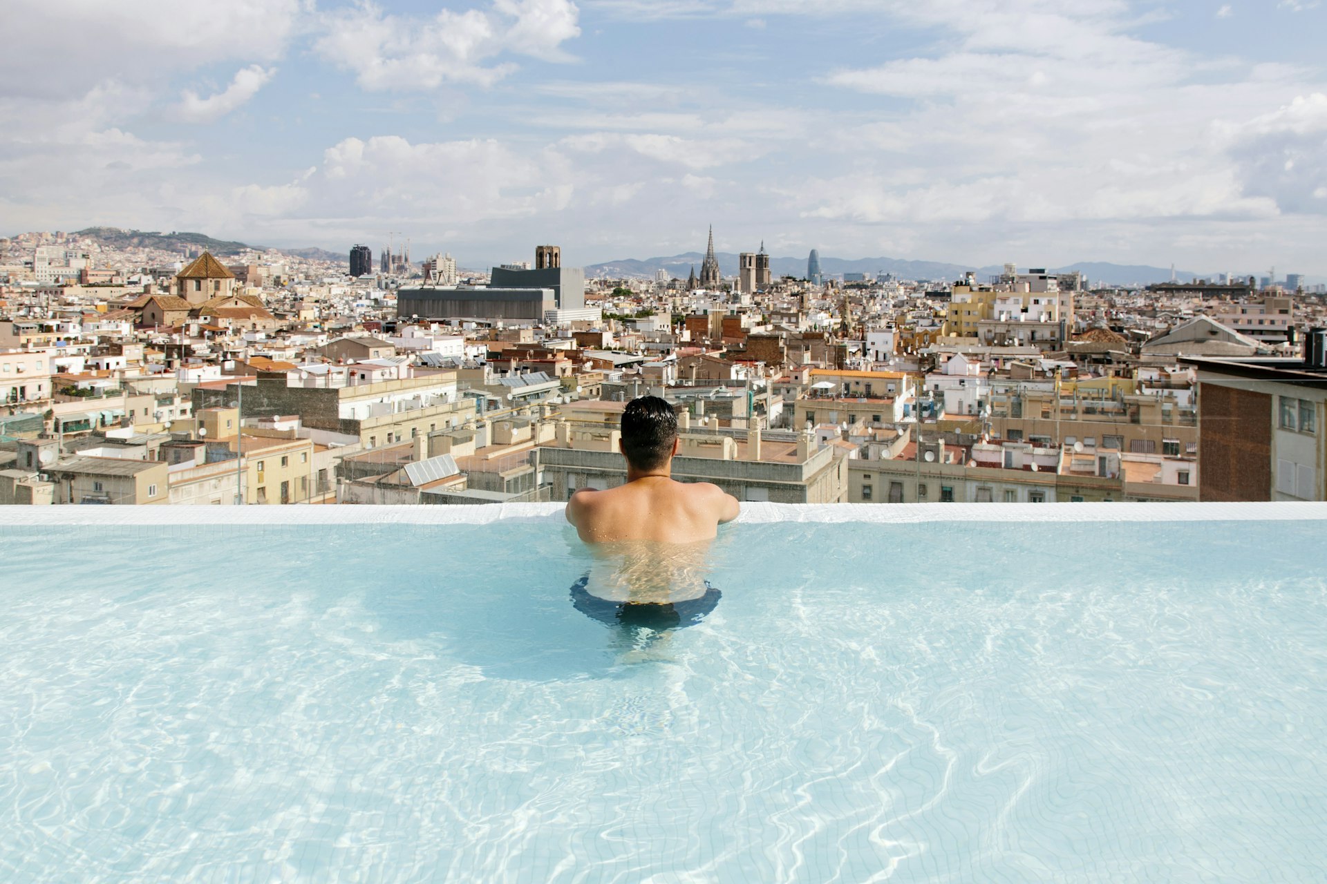 Achteraanzicht van een jonge man die ontspant in het zwembad en kijkt naar de skyline van Barcelona