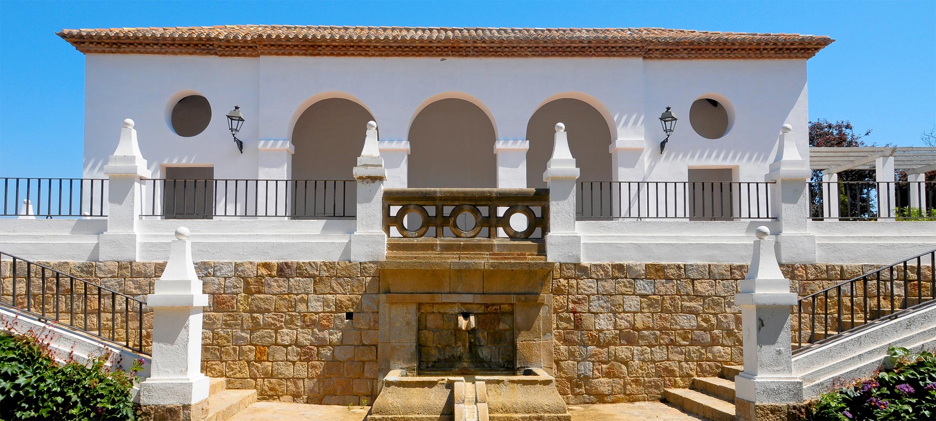 Wit mediterraan gebouw met bogen en fontein.