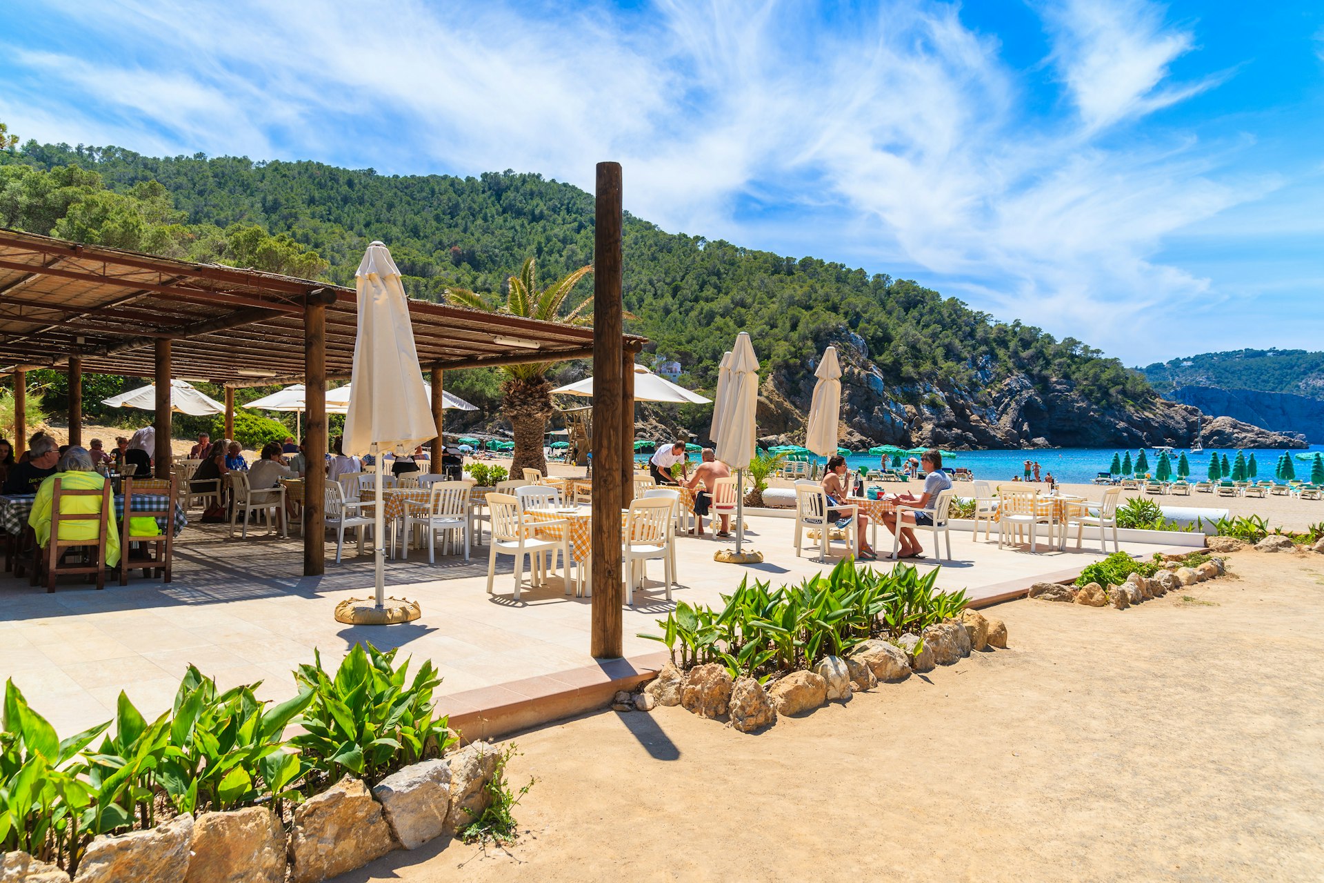 Mensen zitten aan restauranttafels op een terras aan het strand van Cala Benirrás op Ibiza